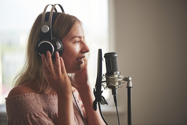Linda cantora loira em fones de ouvido no estúdio de gravação em casa canta uma música em um microfone, ri e sorri de perto