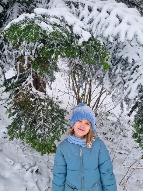 Una linda chica con un abrigo azul bajo un abeto cubierto de nieve