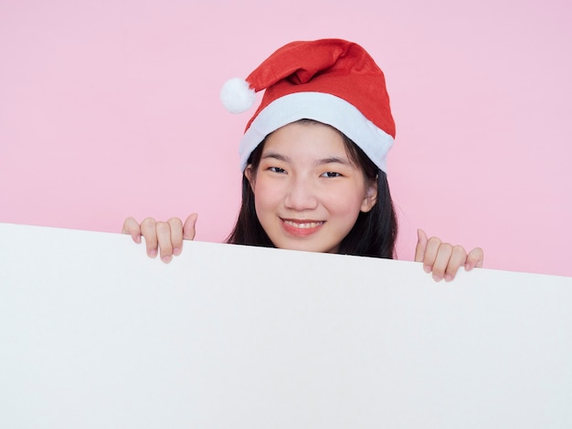 Linda chica adolescente con gorro de Papá Noel con cartel en blanco blanco sobre fondo rosa