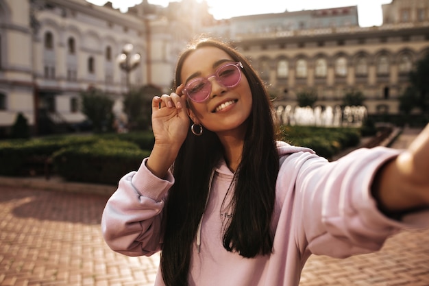 Linda chica adolescente morena con sudadera con capucha rosa y elegantes gafas de sol sonríe sinceramente, mira al frente y toma selfie afuera