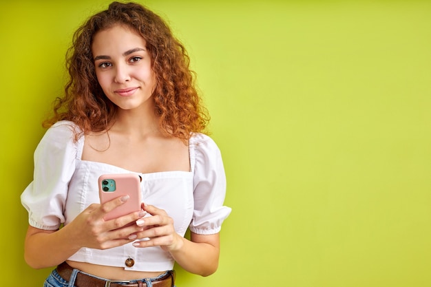 Linda chica adolescente rizada disfruta charlando con alguien que usa teléfono móvil, adorable chica en blusa sostenga el teléfono en las manos, sonríe, aislado en el espacio verde en el estudio