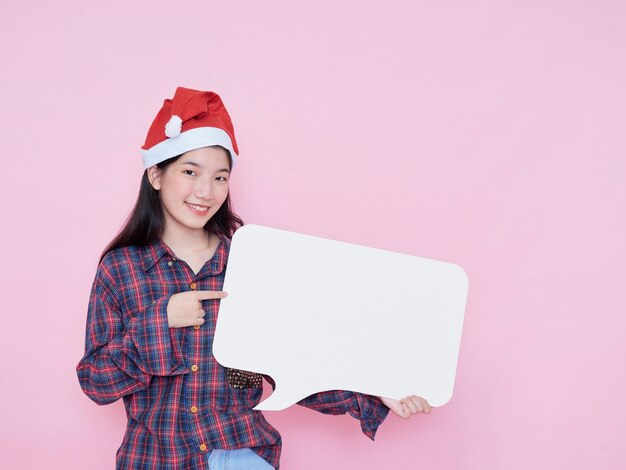 Linda chica adolescente con sombrero de santa que señala el dedo en un cartel en blanco mientras está de pie sobre fondo rosa.