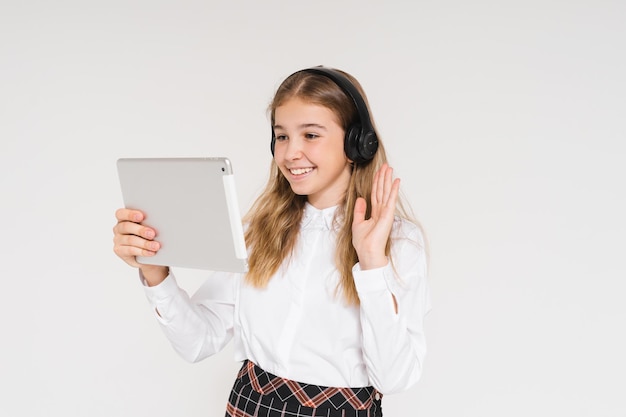 Linda chica adolescente sonriente en uniforme escolar con auriculares y tableta en sus manos aisladas