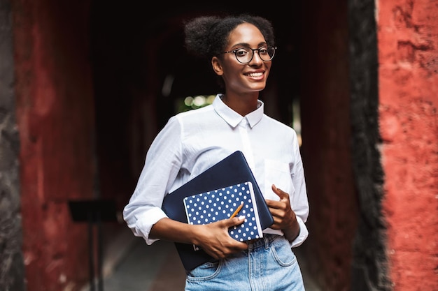 Linda chica africana con anteojos y camisa sosteniendo una computadora portátil y un bloc de notas en las manos mirando alegremente a la cámara al aire libre