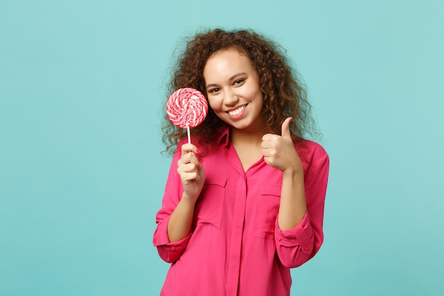 Linda chica africana en ropa casual sosteniendo en la mano piruleta redonda rosa mostrando el pulgar hacia arriba aislado sobre fondo azul turquesa en estudio. Concepto de estilo de vida de emociones sinceras de personas. Simulacros de espacio de copia.