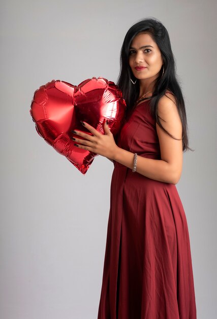 Linda chica alegre en elegante traje rojo posando con globo en forma de corazón sobre fondo gris