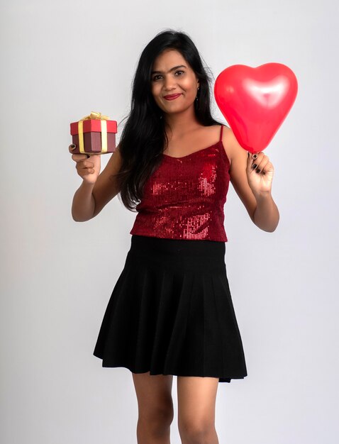 Linda chica alegre en traje elegante posando con caja de regalo y globo en forma de corazón.