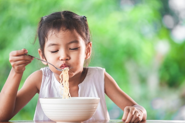 Linda chica asiática niño comiendo deliciosos fideos instantáneos con tenedor
