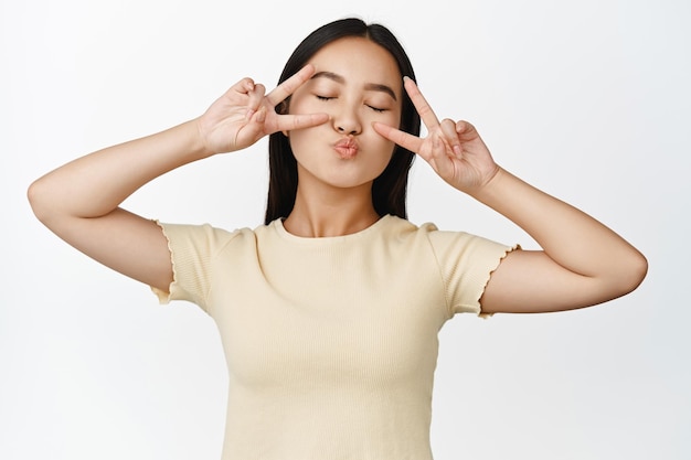 Linda chica asiática soñadora cierra los ojos y los labios fruncidos haciendo cara de beso con signos de paz de pie en camiseta amarilla sobre fondo blanco