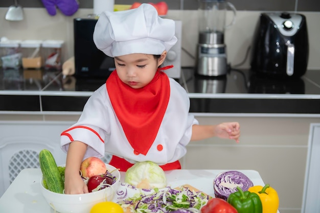 Una linda chica asiática usa uniforme de chef con muchas verduras en la mesa de la cocina. Prepara comida para cenar. Es un momento divertido para los niños.