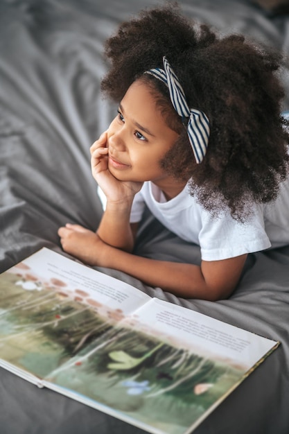 Una linda chica con banda para el cabello acostada en la cama con un libro