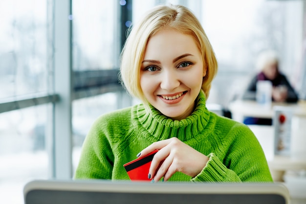 Linda chica con cabello claro con suéter verde sentado en la cafetería con laptop y tarjeta de crédito, retrato, concepto independiente, compras en línea.