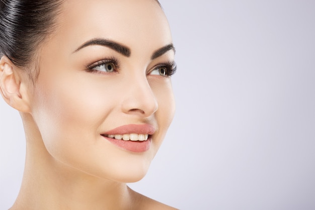 Linda chica con cabello negro fijo detrás, ojos grandes, cejas gruesas y hombros desnudos sosteniendo la mano cerca de la cara en gris, retrato.