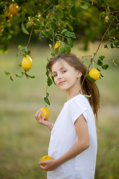 Una linda chica con cabello rubio en una camiseta blanca con limones de verano en el jardín debajo de un árbol