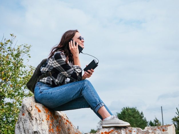 Una linda chica con una camisa a cuadros informal, jeans y gafas está hablando por un teléfono conectado a un banco de energía