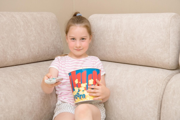 Linda chica en casa en el sofá con un gran vaso de palomitas de maíz cambia los canales de televisión con control remoto