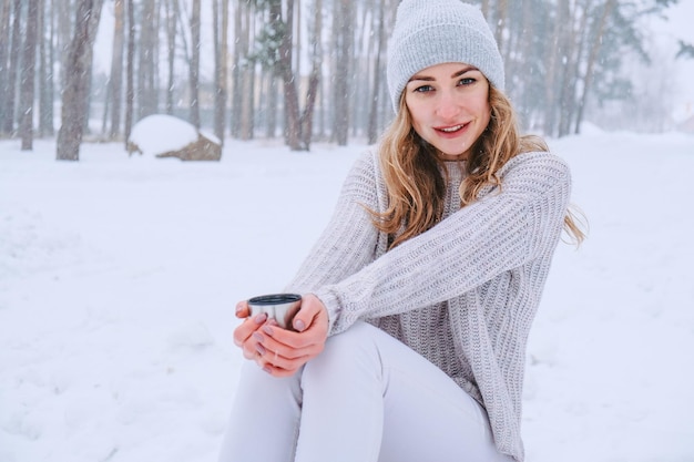 Linda chica caucásica en un suéter blanco y sombrero en un bosque nevado Park bebiendo té caliente o chocolate de un termo. Chica con ropa cálida de invierno durante las vacaciones de Navidad, Año Nuevo