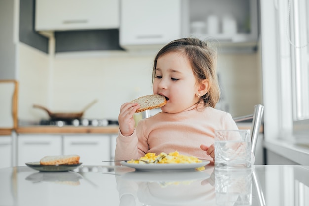 Una linda chica en la cocina está comiendo pan y una tortilla.