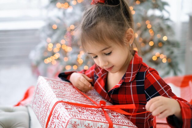 Linda chica desata un lazo rojo en un regalo de Navidad mientras está sentado en una cama junto a un árbol de Navidad