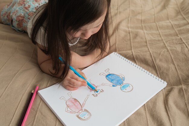 Linda chica dibuja un dibujo a lápiz. Un niño aprende a dibujar personas. El niño se acuesta en la cama y dibuja con un lápiz azul.