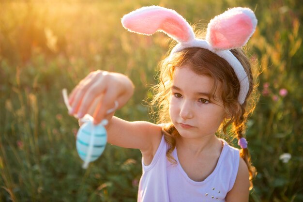 Linda chica divertida con huevos de Pascua pintados en primavera en la naturaleza en un campo con luz solar dorada y flores. Vacaciones de Pascua, conejito de Pascua con orejas, huevos coloridos en una cesta. Estilo de vida