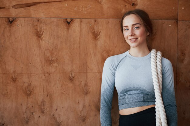 Linda chica está de pie contra la pared de madera y sonriendo. Deportiva joven tiene día de fitness en el gimnasio por la mañana