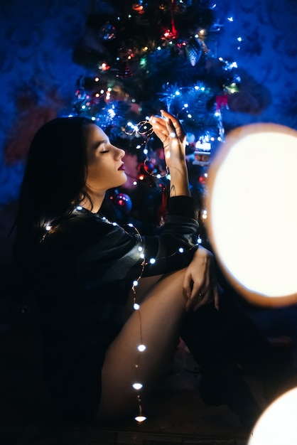 Linda chica está sentada de un festivo árbol de Navidad con una guirnalda