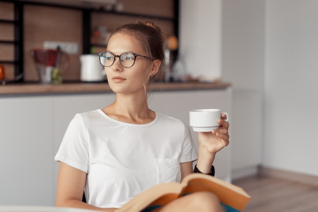 Linda chica está tomando café y leyendo un libro en la mesa de la cocina en casa