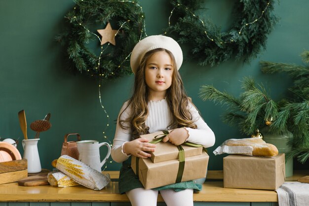 Linda chica con estilo en una boina blanca con regalos de Navidad y sentado en la cocina de color esmeralda, decorada para las vacaciones del Año Nuevo