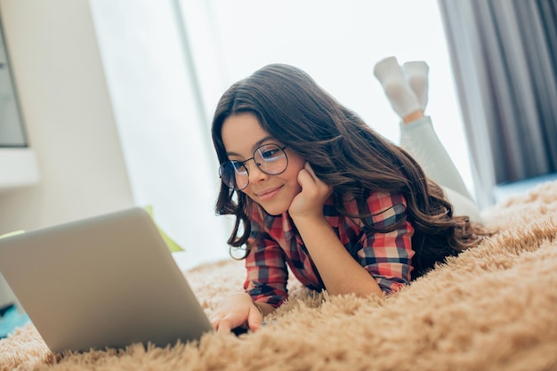 Linda chica con gafas acostada en la cama con el puño tocando la mejilla y sonriendo mientras mira la pantalla del portátil