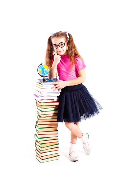 Linda chica con gafas está de pie cerca de la pila de libros con un pequeño globo en la parte superior, aislado sobre fondo blanco.