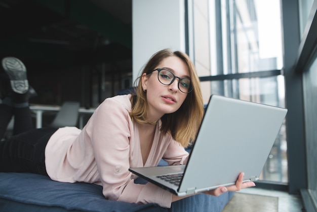 Una linda chica con gafas yace en la ventana del sofá con una computadora portátil en sus manos y mira a la cámara