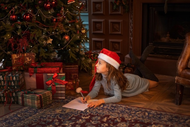 Linda chica con gorro de Papá Noel escribiendo una carta a Papá Noel en la sala de estar decorada de Navidad