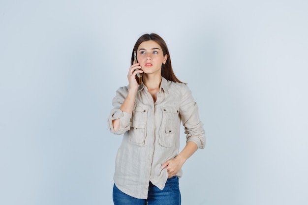 Linda chica hablando por teléfono móvil, mirando a otro lado en camisa y mirando pensativo.