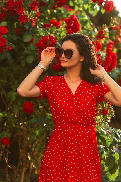 Linda chica hermosa de moda con gafas en un vestido rojo de verano posando cerca de rosas en el parque