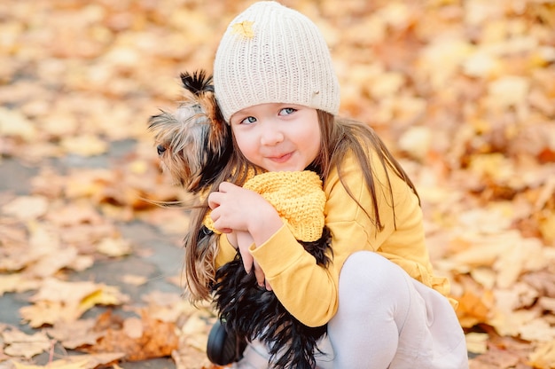 Linda chica con una hoja amarilla caída sobre un sombrero, abraza suavemente a su perro mascota en un parque de otoño con un fondo de hojas amarillas caídas