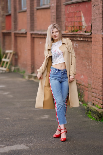 Linda chica en jeans cortos y gabardina caminando por la calle después de la lluvia