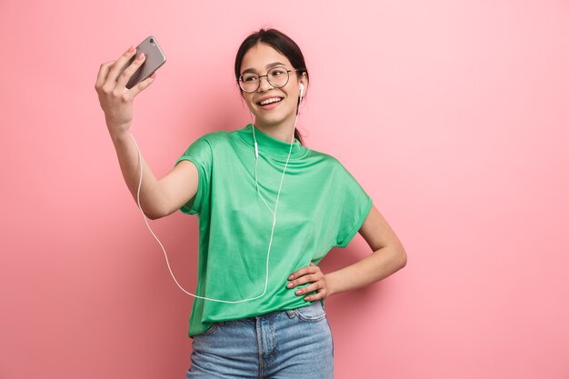 Linda chica joven con anteojos redondos tomando selfie en teléfono móvil mientras usa auriculares aislados sobre pared rosa