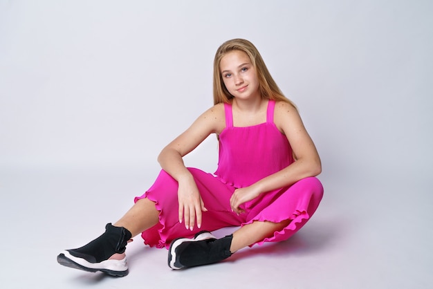 Linda chica joven con cabello largo y rubio de apariencia caucásica posando en un traje rosa sobre una pared blanca sentada con ropa elegante y de moda para un adolescente