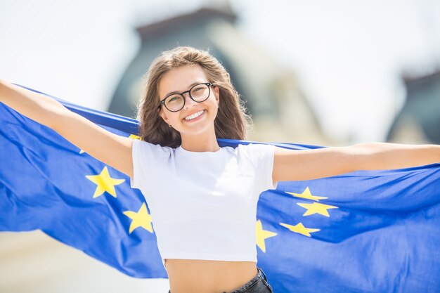 Linda chica joven feliz con la bandera de la Unión Europea frente a un edificio histórico en algún lugar de Europa.