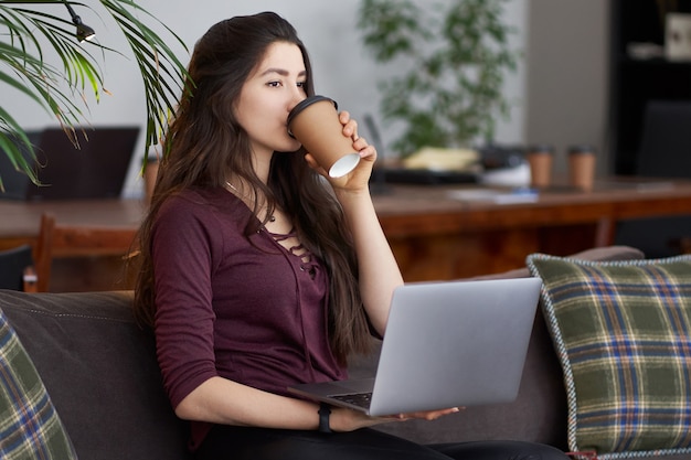 Linda chica joven sentada en el sofá con café y portátil