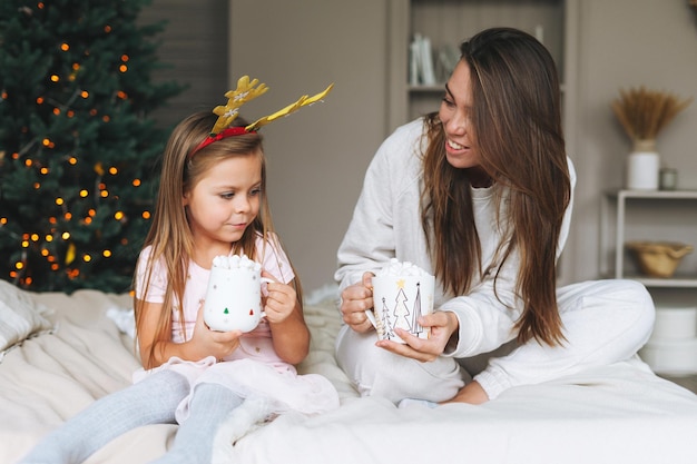 Linda chica con madre en la cama en la habitación con árbol de Navidad Retrato de niño en llanta con cuernos de ciervo