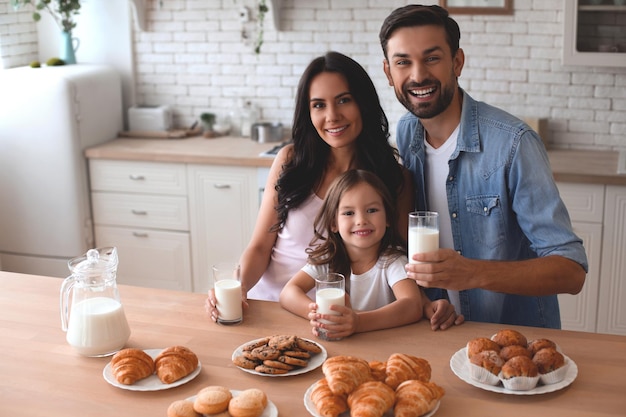 Linda chica madre y padre sosteniendo galletas con vasos de leche y mirando a la cámara y en la cocina en casa