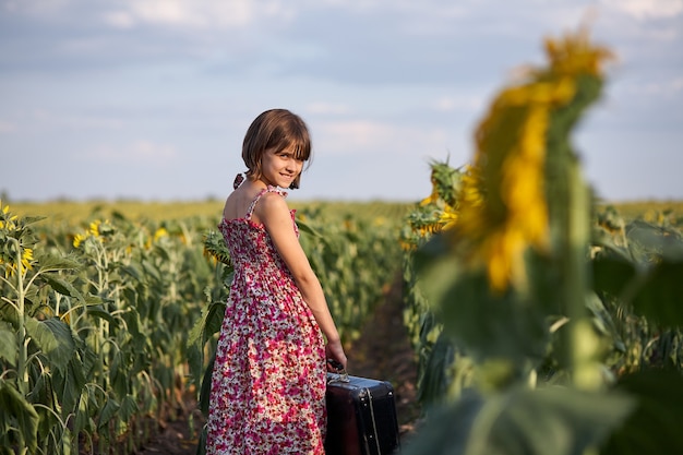 Linda chica con maleta vieja en un campo de girasoles.