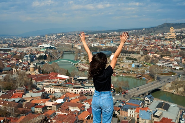 Una linda chica morena disfruta del impresionante paisaje de Tbilisi desde la colina. Toda la ciudad a sus pies.