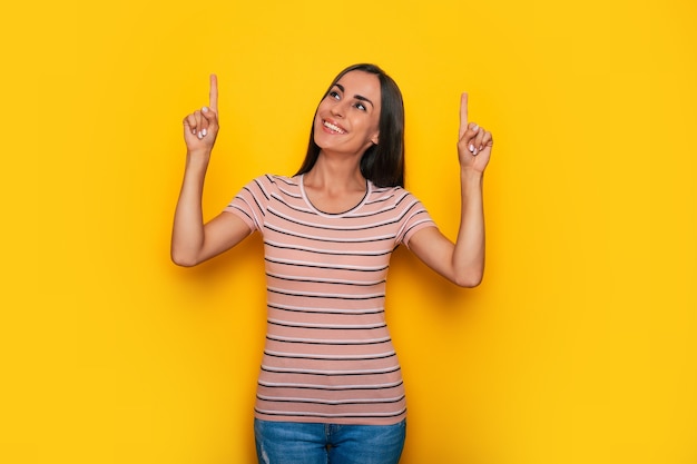 Foto linda chica morena joven sonriente emocionada está apuntando hacia fuera para algún anuncio mientras se encuentra en el fondo de la pared amarilla