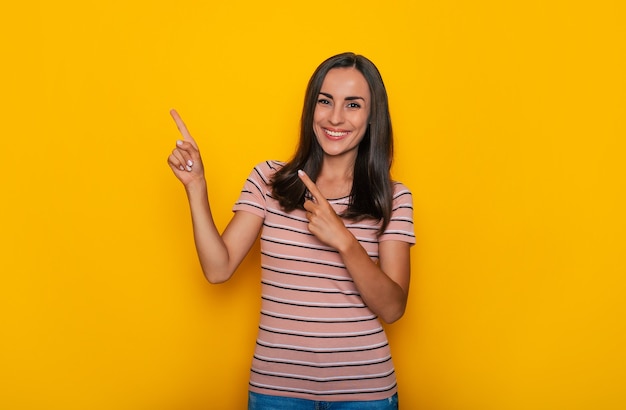 Linda chica morena joven sonriente emocionada está apuntando hacia fuera para algún anuncio mientras se encuentra en el fondo de la pared amarilla