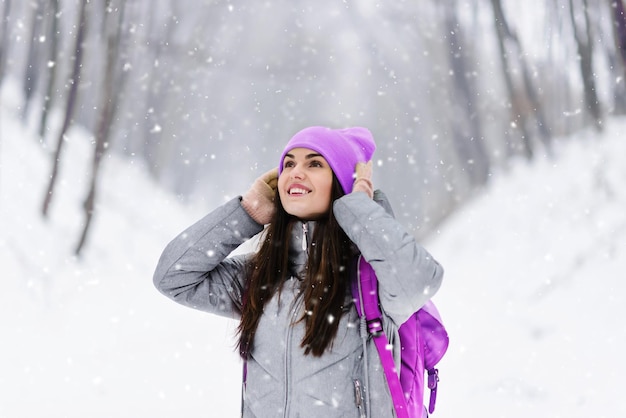 Linda chica morena con mochila con sombrero morado y chaqueta gris se regocija de la nieve mientras camina en el bosque nevado