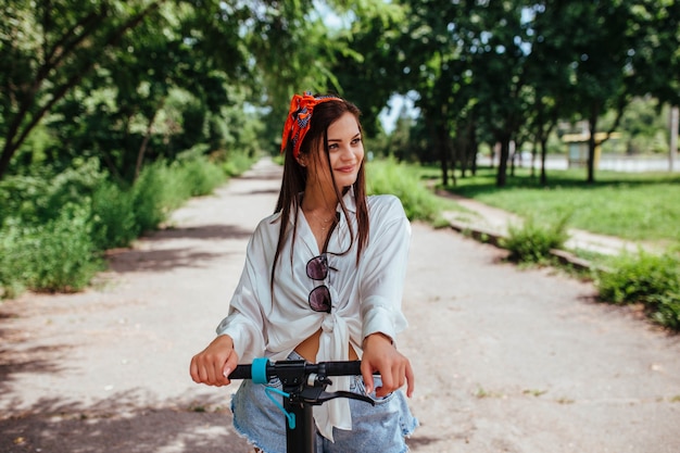 Linda chica morena monta en un scooter de electrodos en el parque que lleva una camisa blanca. concepto de transporte ecológico y alquiler.