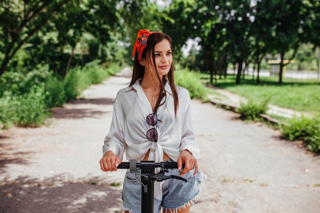 Linda chica morena monta en un scooter de electrodos en el parque que lleva una camisa blanca. concepto de transporte ecológico y alquiler.